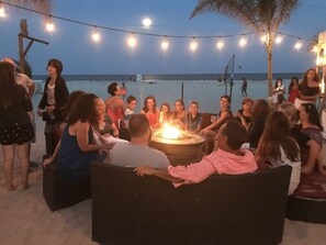 A group of people gathered around a fire pit on a beach at dusk, with string lights overhead.