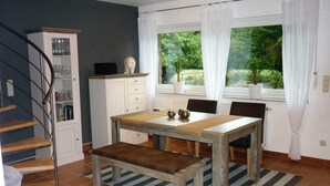 A dining area with a wooden table, bench, and chairs. There is a cabinet with glassware and a potted plant by the window. - Holiday house in the green north of the Ruhr area (Dinslaken)