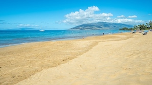 Una extensa playa arenosa con aguas azules cristalinas, un velero a lo lejos y una montaña al fondo.