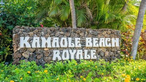 Un letrero de piedra con la inscripción "KANOLE BEACH ROYALE", rodeado de exuberante vegetación y flores.