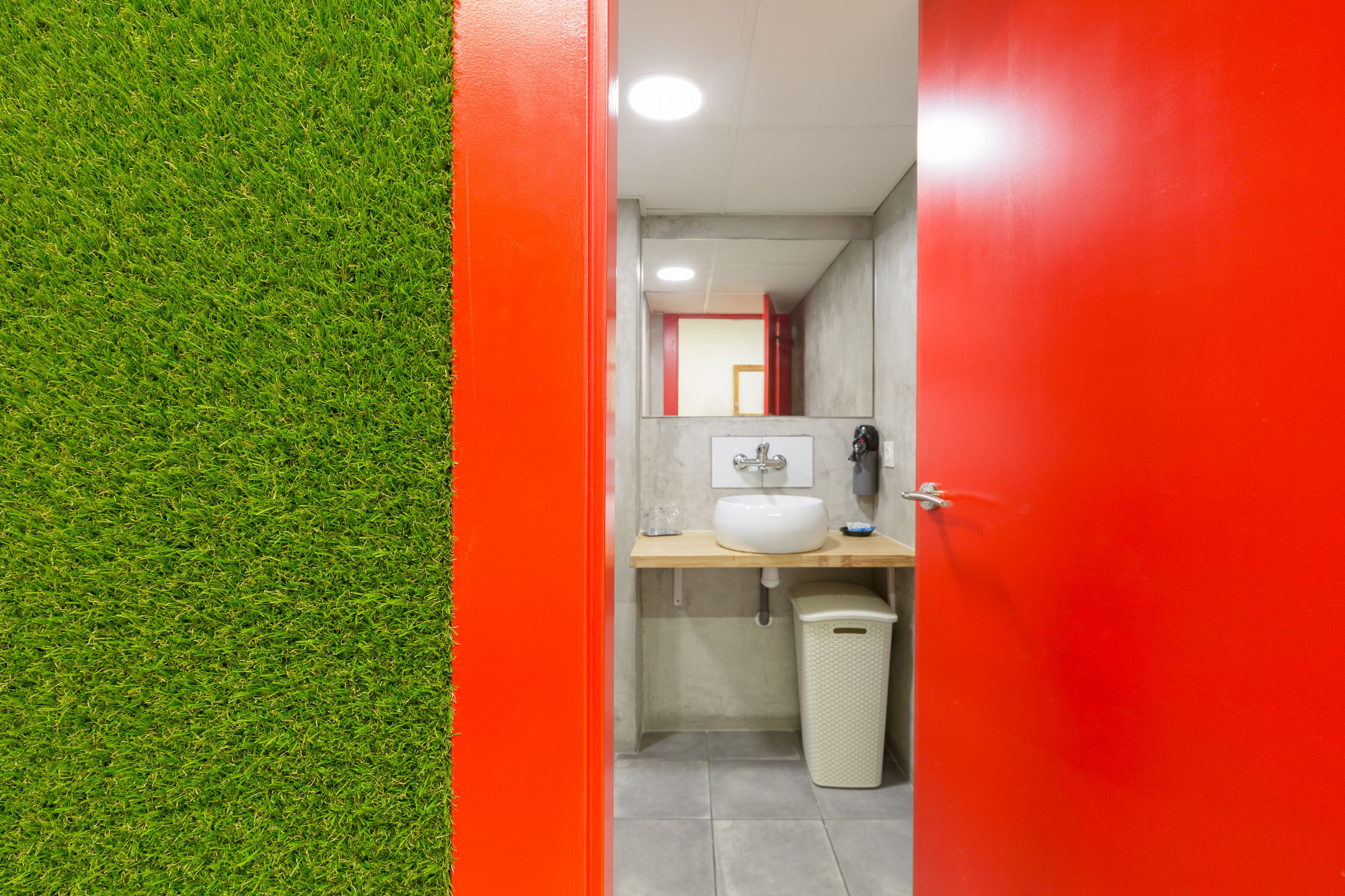 Un baño con pared de césped artificial verde, un lavamanos blanco, un espejo y un cubo de basura.