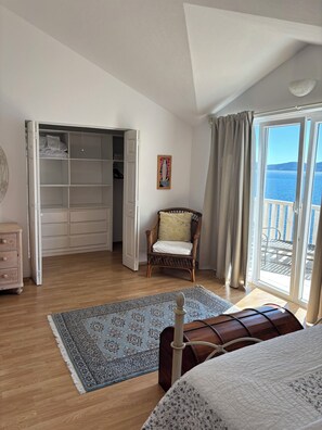 A bedroom with a wooden floor, a bed with a white quilt, a wicker chair, a wardrobe, and a view of the sea through a balcony. - Enjoy Your Front Row Villa On The Adriatic Sea (Pisak)