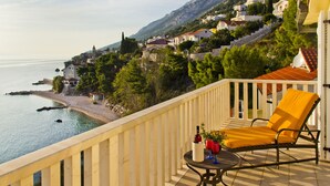 A balcony with a yellow chair, a table with a bottle and glasses, overlooking a coastal town with houses and a mountain in the background. - Enjoy Your Front Row Villa On The Adriatic Sea (Pisak)