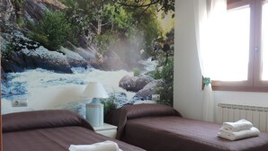 A hotel room with two beds, a mural of a river, a lamp, and a window with curtains.