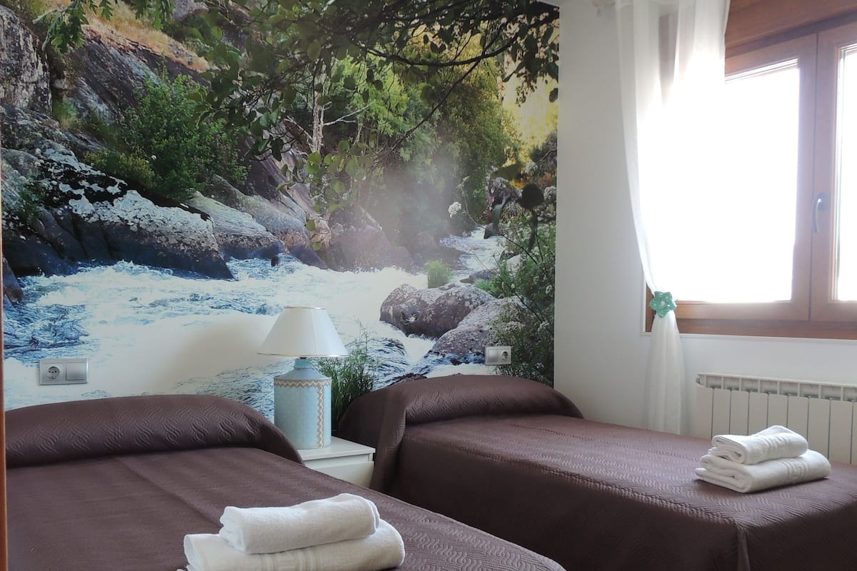 Habitación de hotel con dos camas, un mural de un río, una lámpara y una ventana con cortinas.