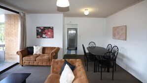 En stue med en brun sofa, et spisebord med stole og et køleskab i baggrunden.