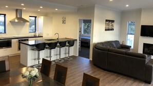 A modern kitchen with a central island, bar stools, and a dining table with chairs. There is a living area with a sofa and a flat-screen TV.
