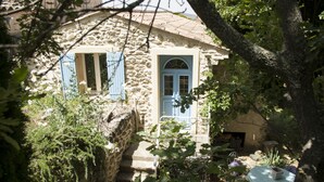 A stone building with a blue door and window shutters, surrounded by greenery and a small table with a bowl.