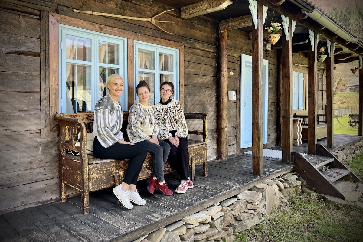 Trois personnes sont assises sur un banc en bois devant une cabane rustique, qui possède des marches en pierre et une véranda en bois.