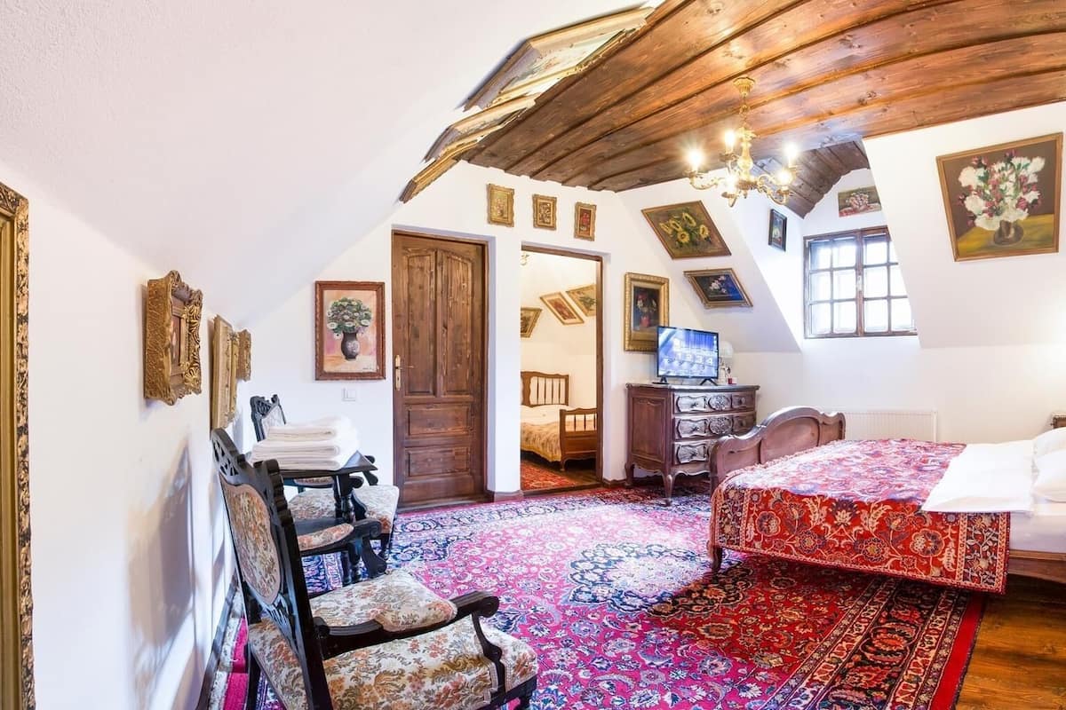 Une chambre traditionnelle avec un plafond en pente, des poutres apparentes, un lit recouvert d’une courtepointe à motifs rouges, une commode en bois et une chaise agrémentée d’un coussin à motifs.