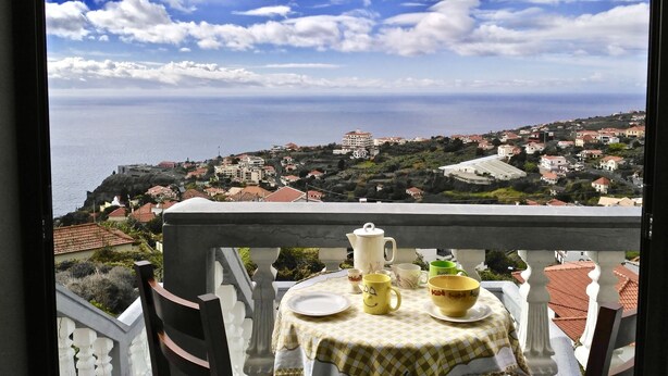 A balcony with a table set for tea, overlooking a coastal town with buildings and the sea.
