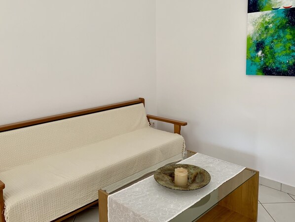 Une pièce avec un canapé en bois adossé à un mur blanc, une petite table avec une bougie et un tableau représentant une scène abstraite aux couleurs bleue et verte.
