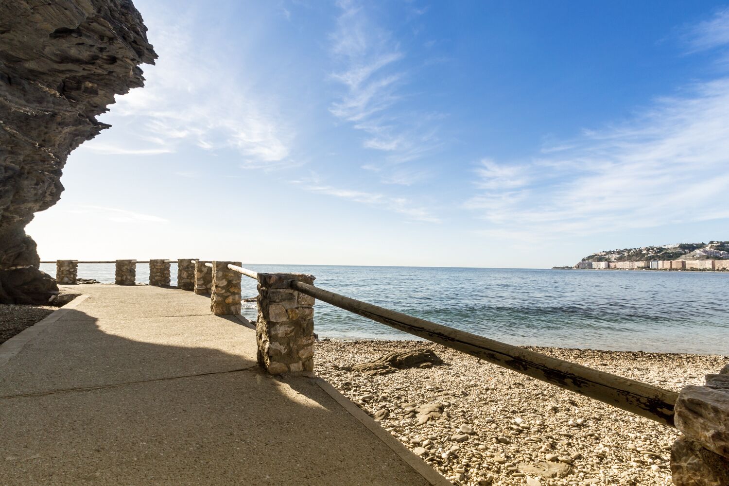 Un paseo marítimo con pilares de piedra, un camino de concreto y vista al mar y una colina distante.