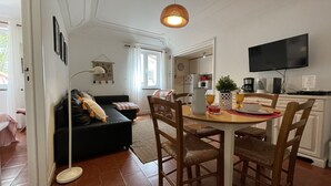 Un salon avec un canapé noir, une table à manger avec des chaises, un téléviseur et, au fond, un coin cuisine.