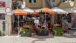 Un café situé au bord de la rue, avec des tables en terrasse, un tableau affichant le menu et un magasin à proximité.