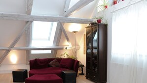 Ein Loft-Zimmer mit freiliegenden Holzbalken, einem roten Sofa, einem dunklen Holzschrank und einem großen Fenster mit Tageslicht.