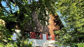 Ein Haus mit weißer Fassade und roten Fensterläden, umgeben von üppiger Vegetation und einem Schubkarren voller gelber Blumen.