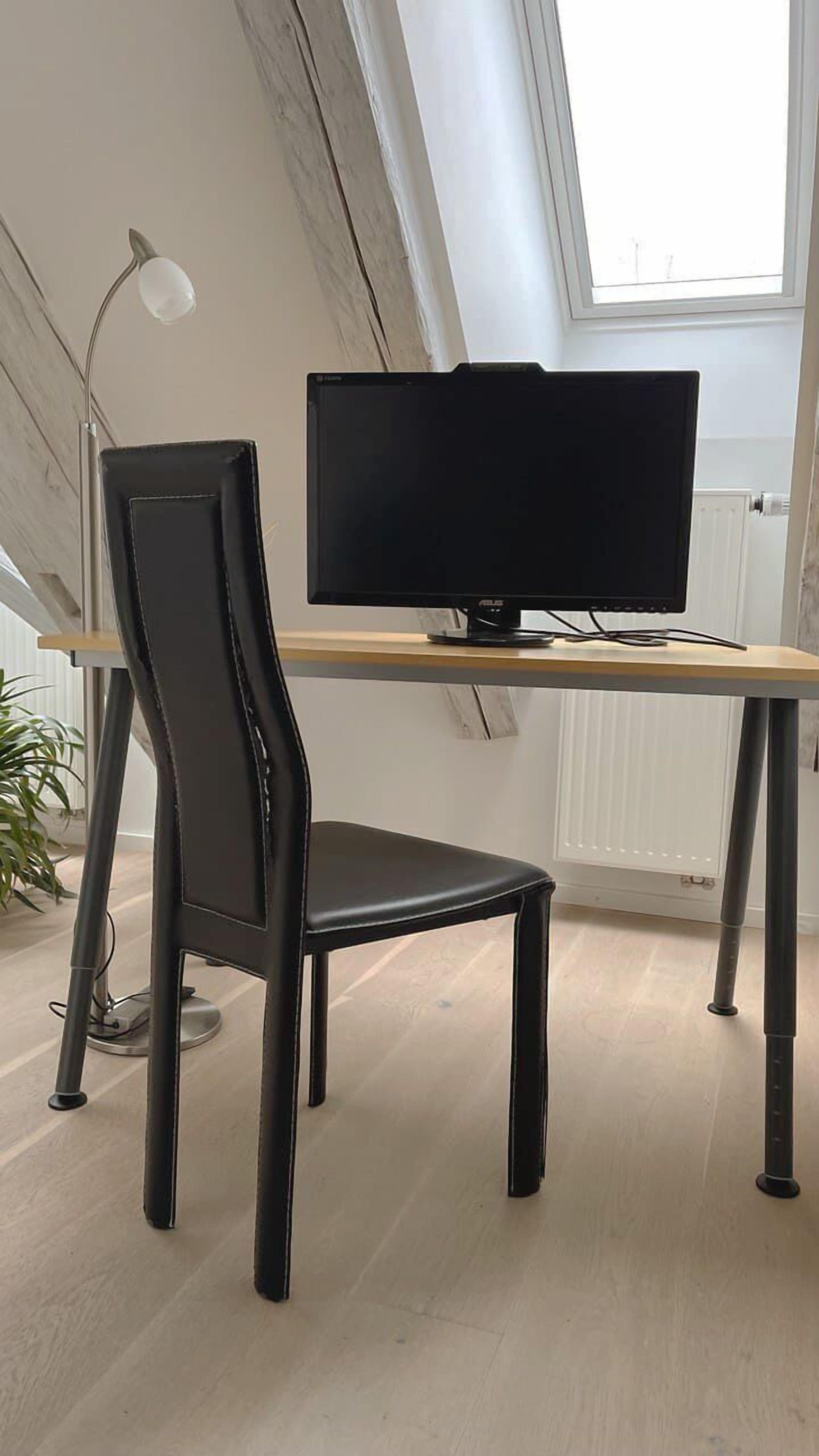 Una silla de oficina moderna y un monitor de pantalla plana sobre un escritorio de madera en una habitación con techo inclinado y una ventana.