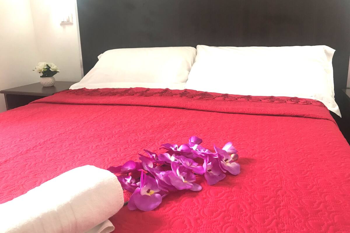 A bed with a red bedspread, white pillows, and a bouquet of purple flowers.
