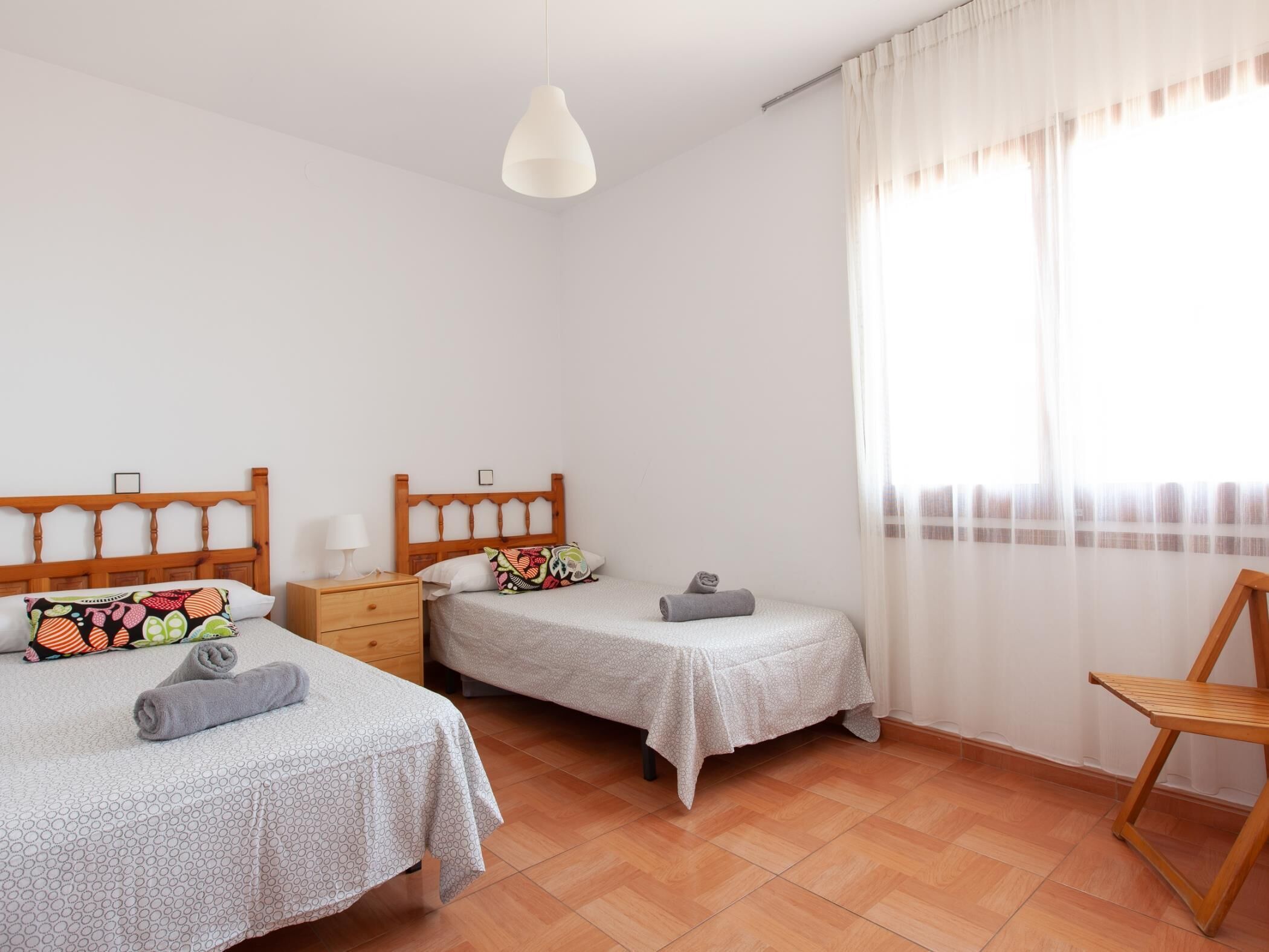 Une chambre avec deux lits, une chaise en bois et une fenêtre ornée de rideaux légers.