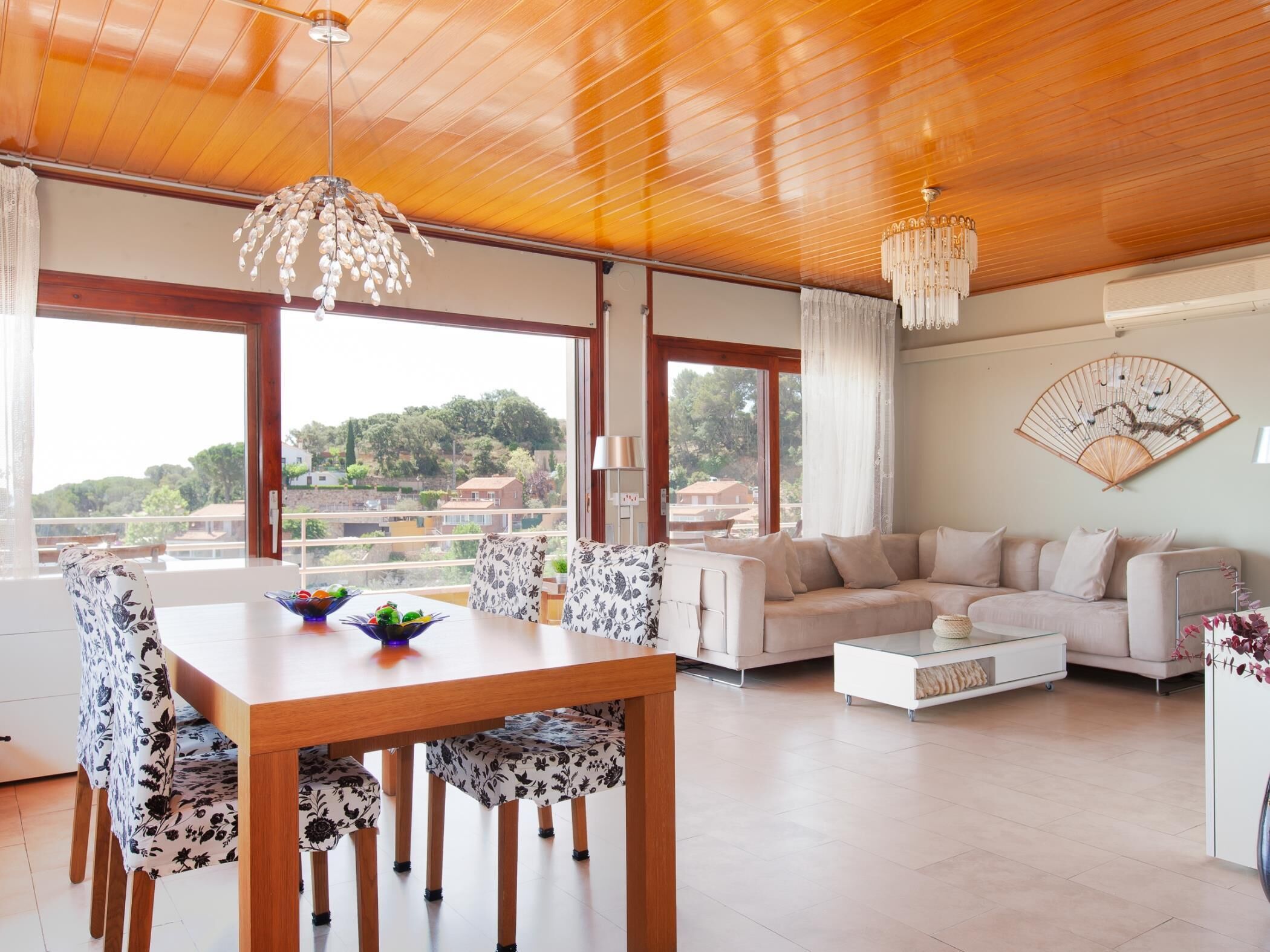 Un salon spacieux doté d’un plafond en bois, un coin repas avec des chaises à motifs floraux, un canapé beige et une grande fenêtre offrant une vue sur le paysage.