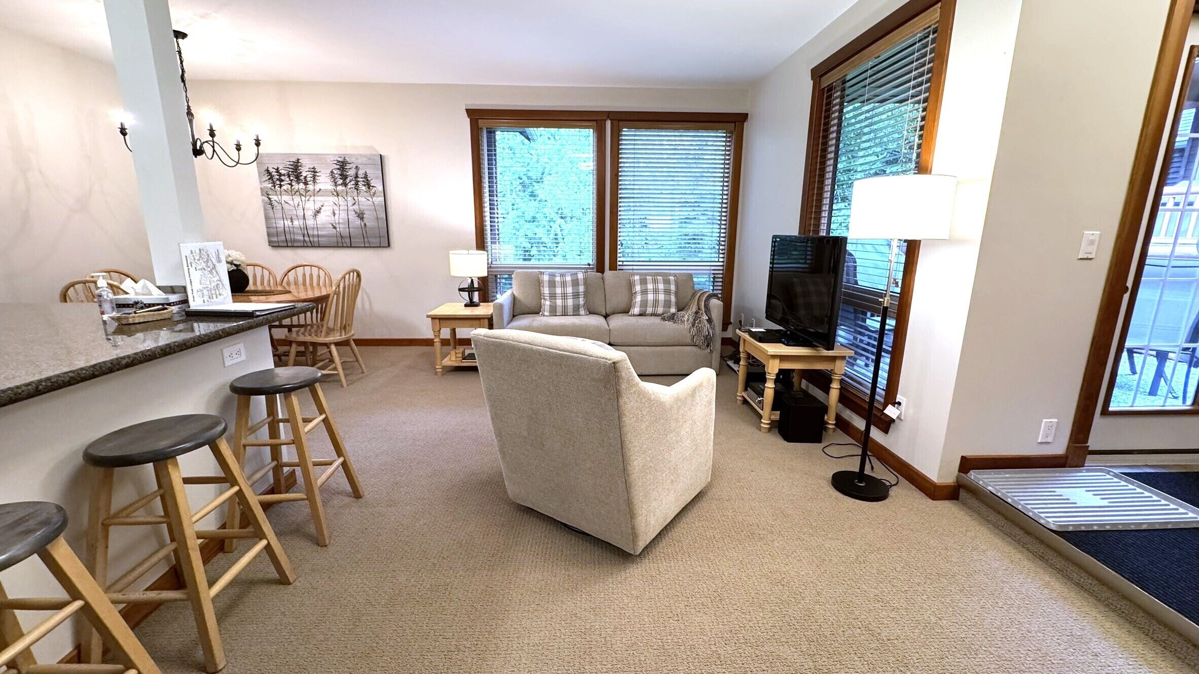 A living room with a sofa, a TV on a stand, a dining area with chairs and a table, and a kitchen area with a counter and stools.