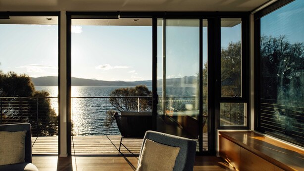 Una habitación moderna con amplios ventanales que dan a un lago, una terraza de madera y una zona de estar con una silla y un sofá.