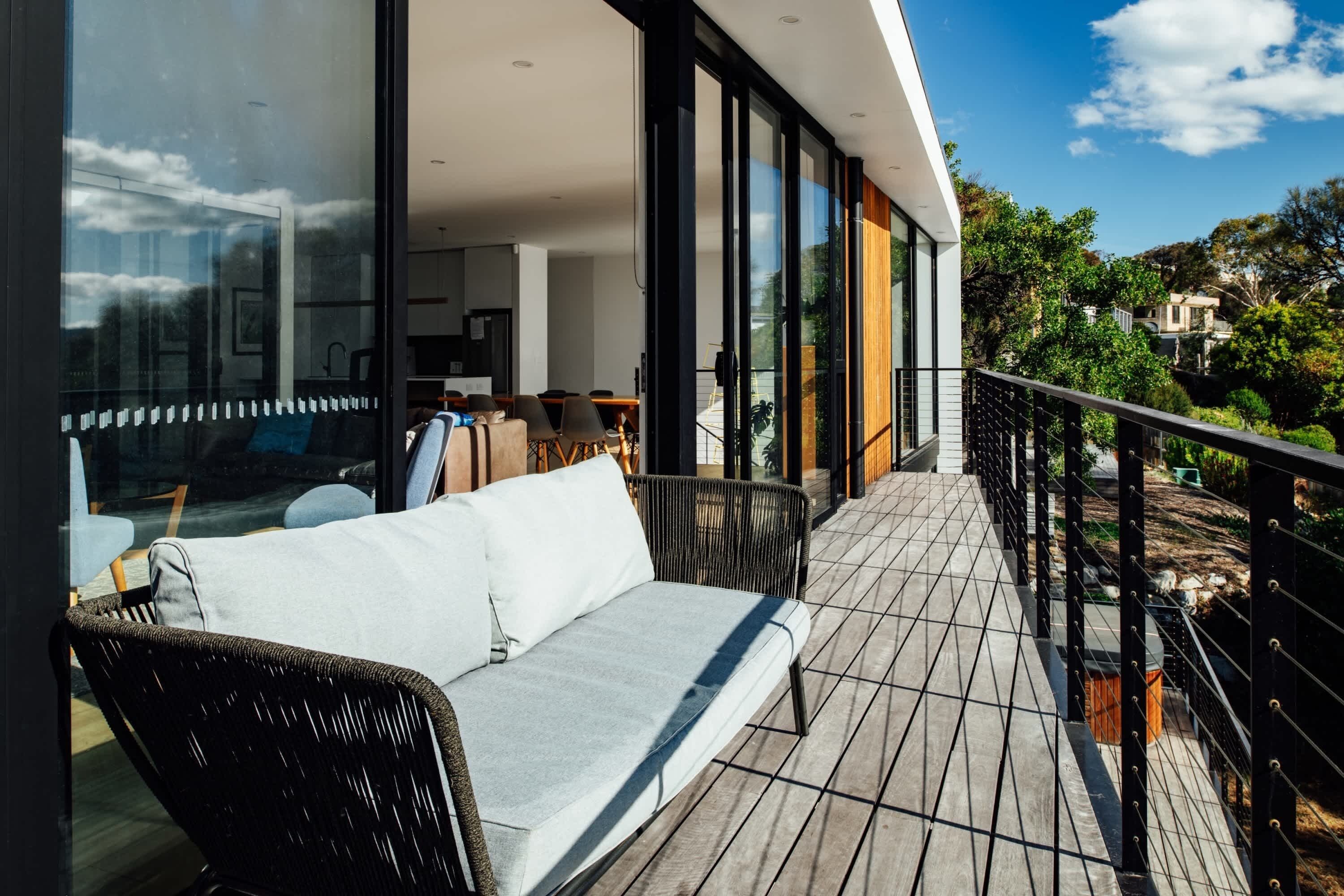 En moderne balkon med møbler, heriblandt en sofa og fletstole, store glasdøre der fører til et spiseområde, samt udsigt over det omkringliggende landskab.