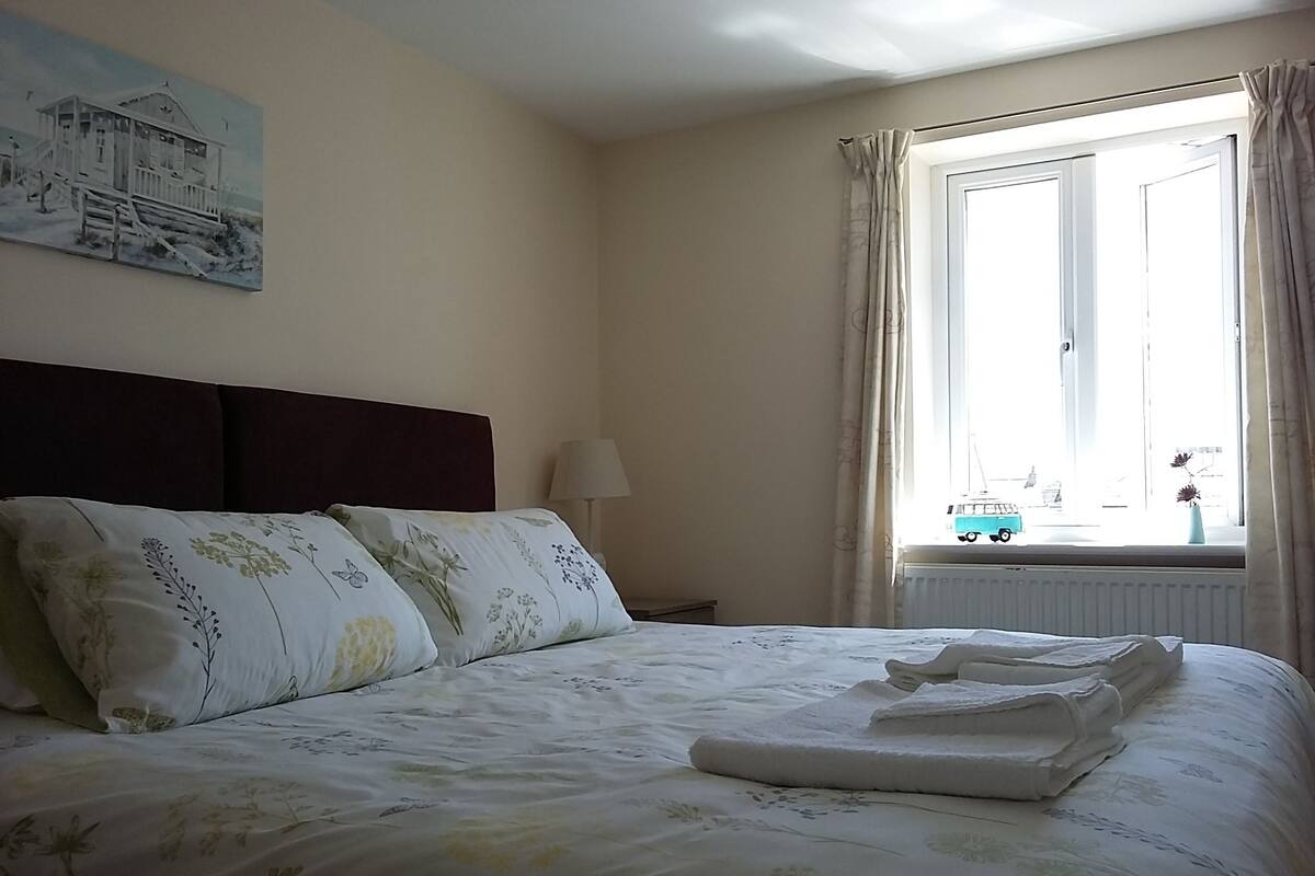 A neatly made bed with floral bedding, a headboard, a lamp, and a window with curtains.