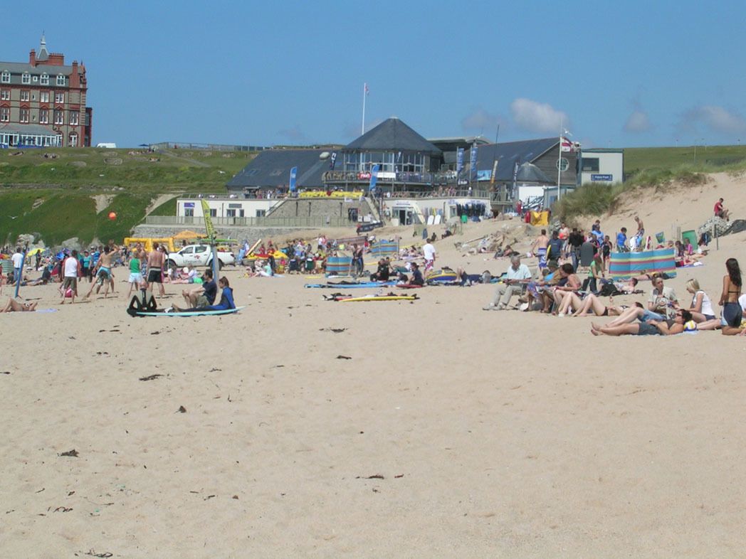 A beach with people sunbathing and swimming, a lifeguard tower, and a building with a sloped roof.