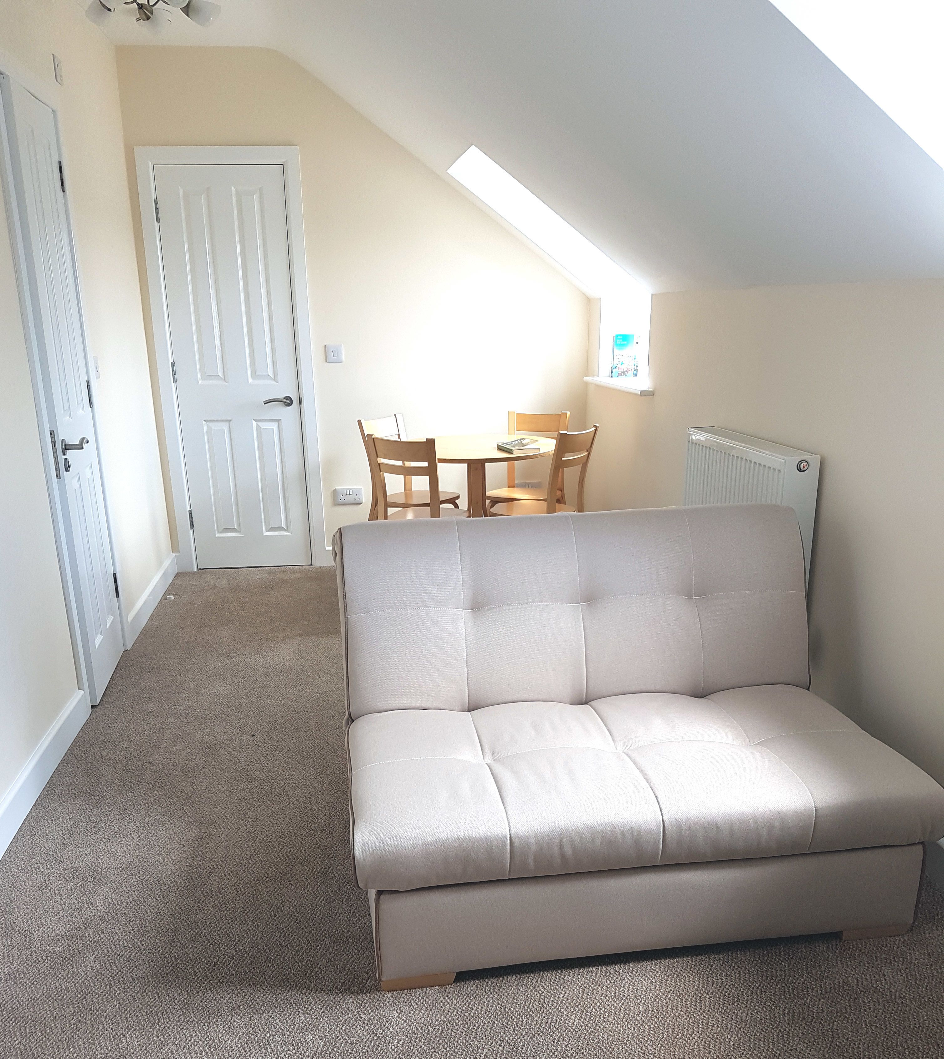 A small, well-lit room with a beige sofa, a dining table with chairs, and a white door.