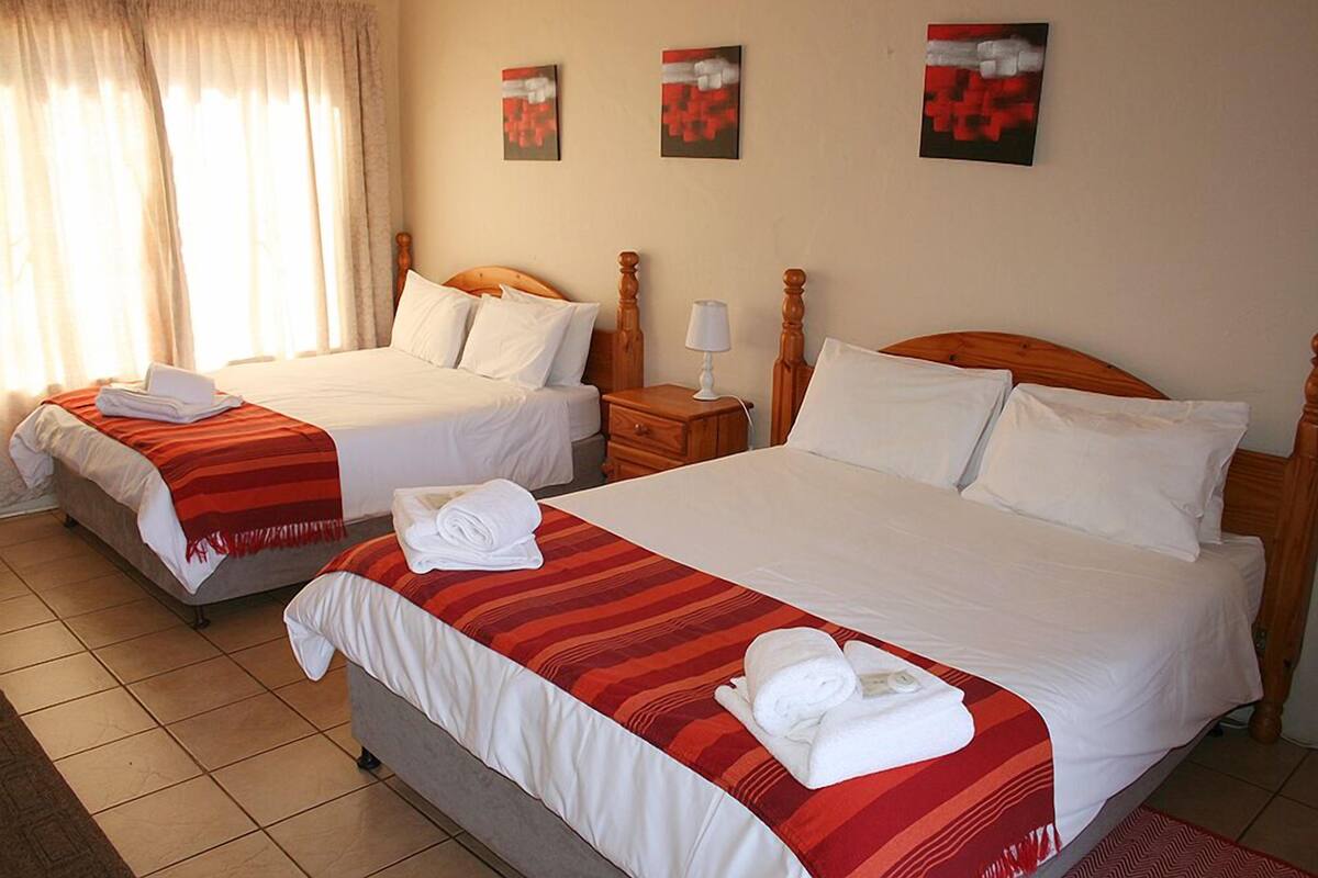 Une chambre d’hôtel avec deux lits, chacun recouvert de draps blancs et d’une couvre-lit à rayures rouges. Trois tableaux ornent le mur, il y a une table de chevet avec une lampe, et une fenêtre avec des rideaux.