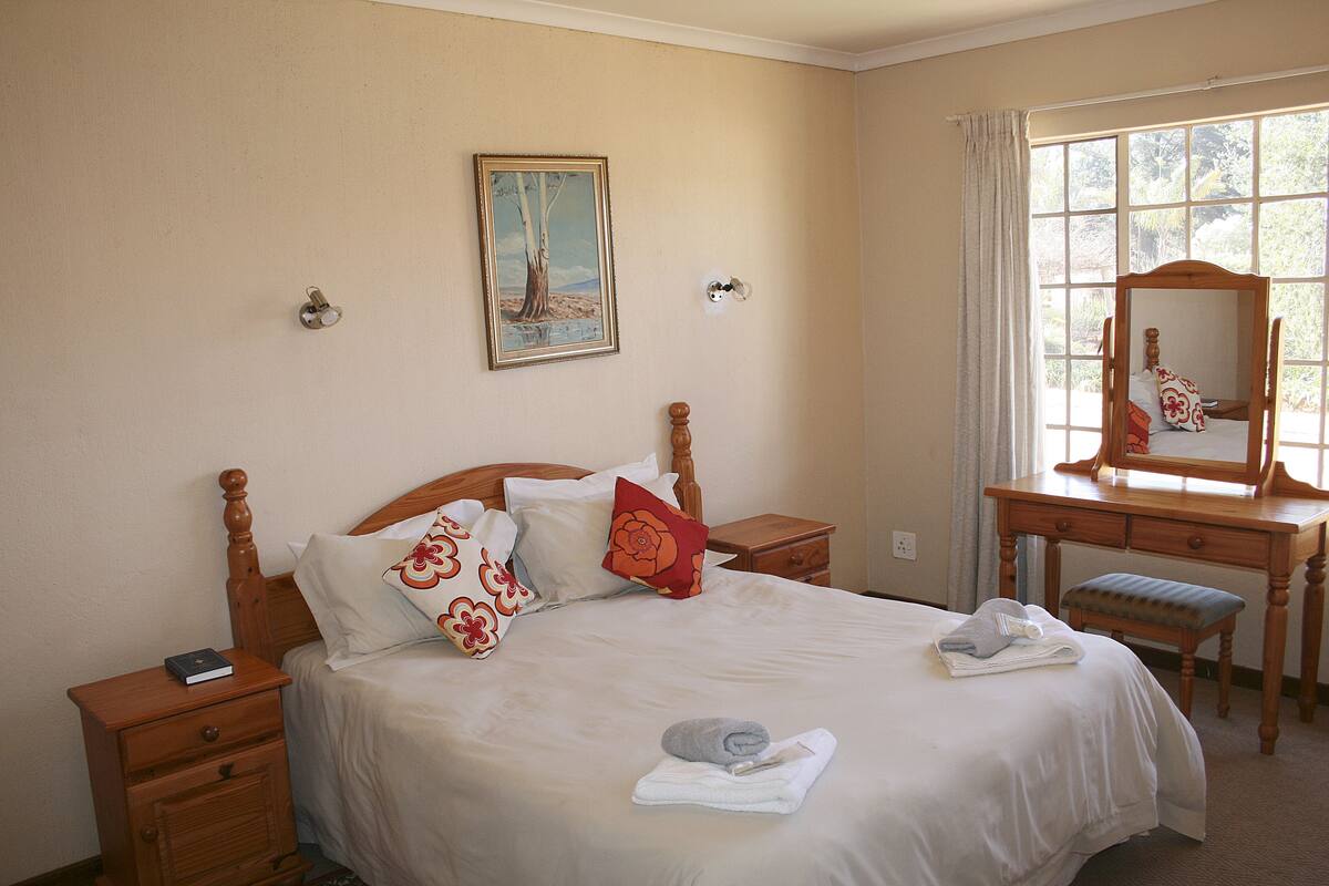 Un lit bien fait, avec des oreillers, une table de chevet, une commode en bois avec un miroir et une fenêtre avec des rideaux.