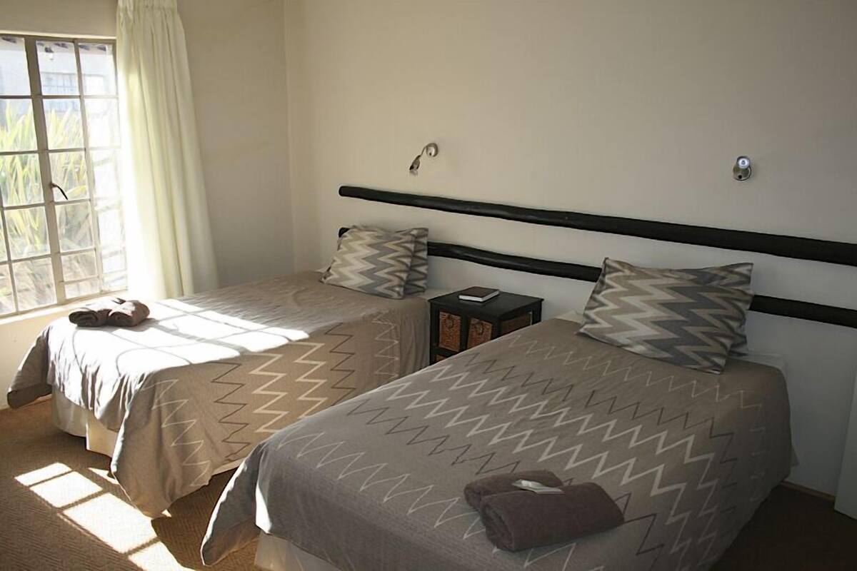 Une chambre avec deux lits, chacun doté d’une tête de lit et d’oreillers, une petite table avec un livre, et une fenêtre avec des rideaux.