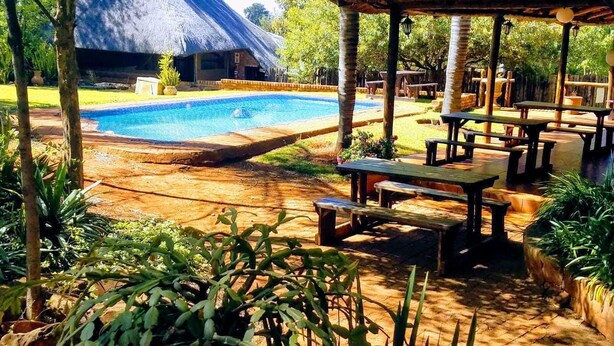 A pool area with a thatched-roof building, wooden picnic tables, and surrounding greenery.