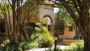 A courtyard with a building featuring an arched entrance, surrounded by lush greenery and a water feature.