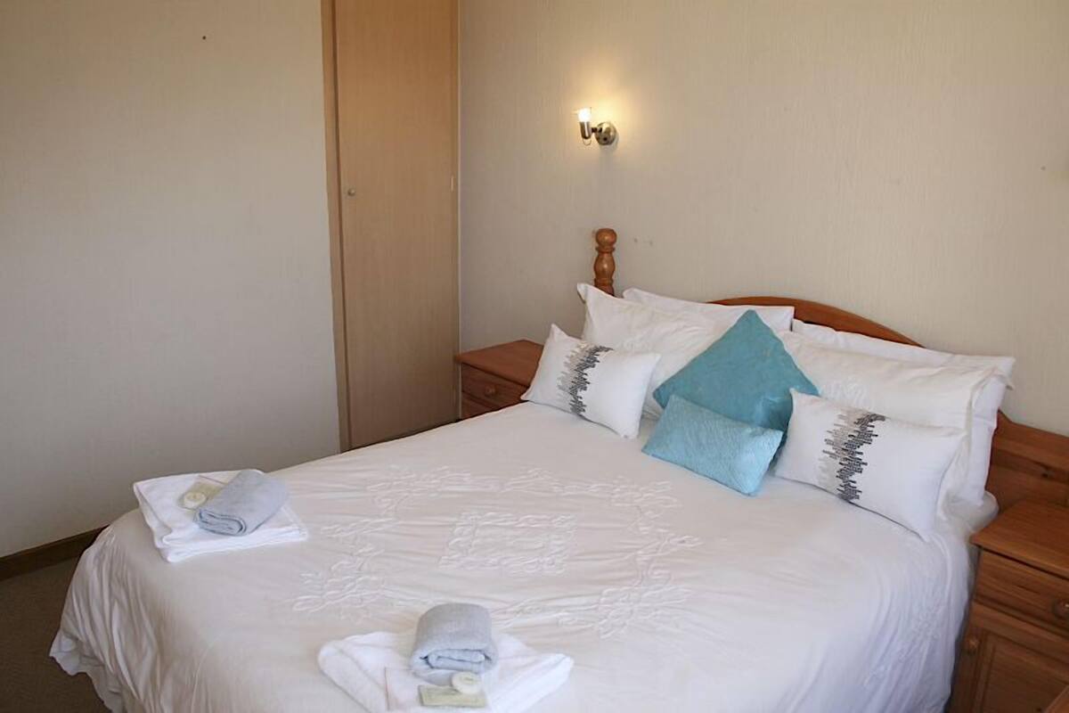 Un lit bien fait, avec du linge de lit blanc et plusieurs oreillers, dont un bleu, dans une chambre sobre, dotée d’une table de chevet en bois et d’un luminaire fixé au mur.