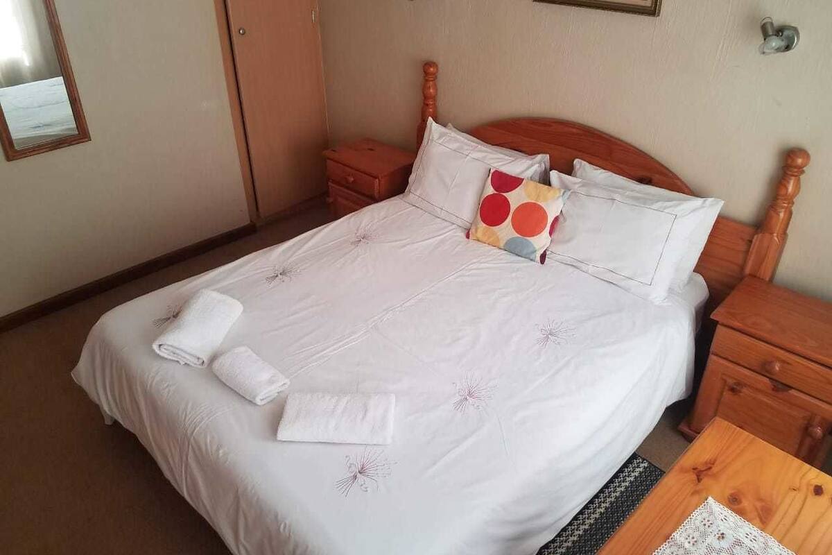 Un lit bien fait, avec du linge de lit blanc orné d’un motif floral, une table de chevet en bois avec un tiroir, et une petite table en bois sur laquelle est posée une nappe.