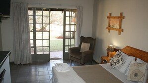 A hotel room with a bed, a chair, a small table with a lamp, and a view through a door to an outdoor area.