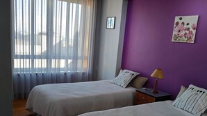 Una habitación con dos camas, una ventana con cortinas, una pared morada y una pequeña mesa con lámpara.