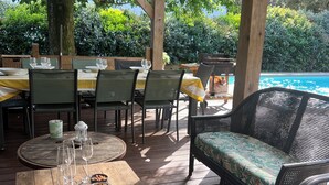 Area pranzo all'aperto con tavoli e sedie in legno, una piscina sullo sfondo e luci decorative appese.