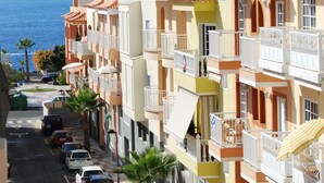 Eine Reihe farbenfroher Gebäude im mediterranen Stil mit Balkonen, eine Straße mit parkenden Autos und Blick auf das Meer.