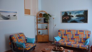 Uma sala de estar com dois sofás coloridos, uma mesa de centro com livros e uma tigela de frutas, uma estante de vime com plantas e quadros nas paredes.