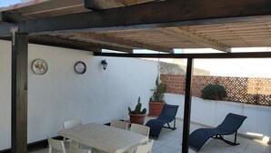 Um alpendre coberto com mesa e cadeiras, plantas em vasos e uma cadeira de descanso azul.