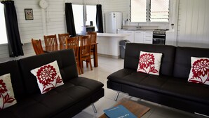 A modern living room with a black sofa, a dining table with chairs, and a kitchen area with white cabinets and appliances. - Mac's Shack - Titikaveka (Titikaveka)