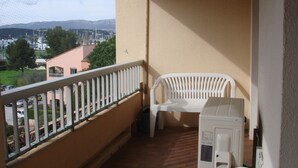 Un balcon avec une chaise en plastique blanche et un lave-linge, donnant sur un quartier résidentiel avec des maisons et une montagne visible au loin.