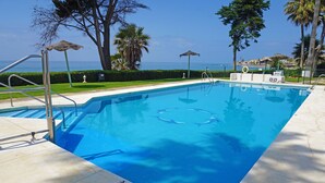 A clear blue pool with a view of the ocean, surrounded by palm trees and a well-maintained lawn. - Beachfront apartment with amazing views & newly renovated with WiFi and int. TVs (Mijas)