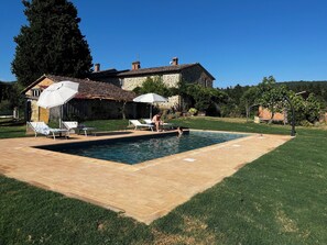 A swimming pool with lounge chairs and umbrellas, surrounded by a well-maintained lawn and a stone building in the background.