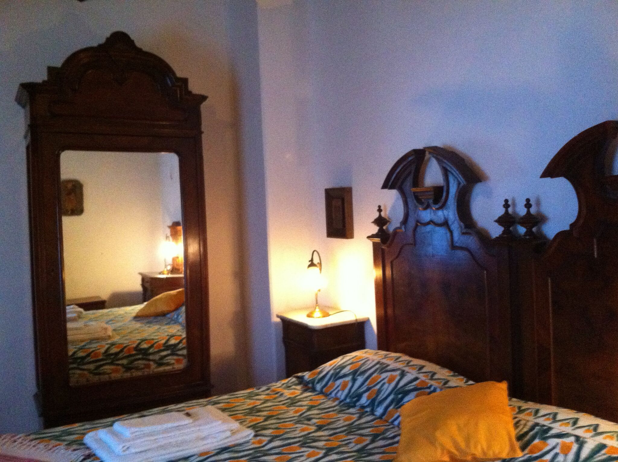 A bedroom with a large mirror, a wooden headboard, a bedside table with a lamp, and a bed with patterned bedding.