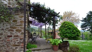 Ein Steinhaus mit einer hölzernen Pergola, die mit violetten Blüten bewachsen ist, einem Blumentopf und einem kleinen Garten mit weißen Blumen.